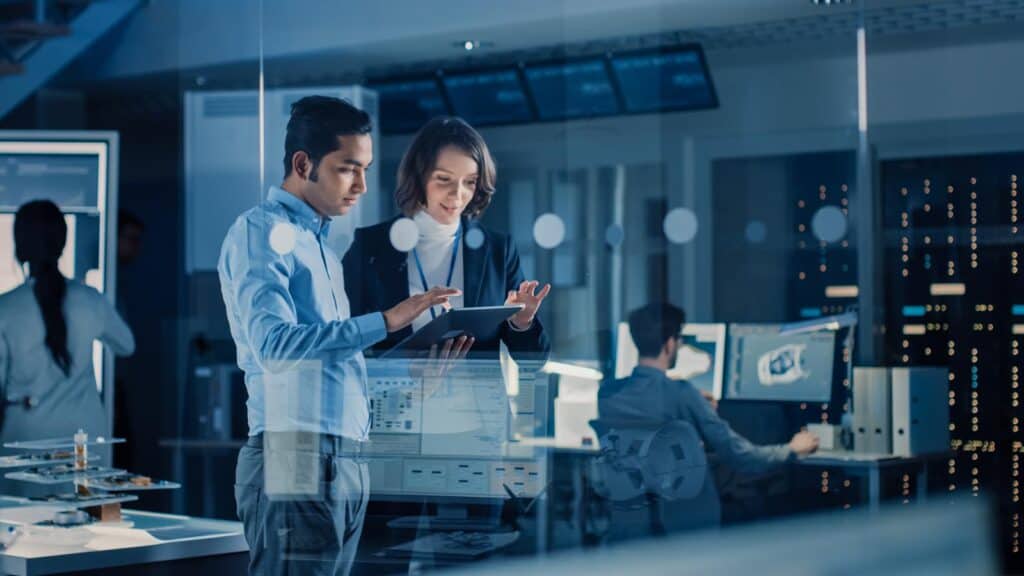 In a Technology Research Facility: Female Project Manager Talks With Chief Engineer, they Consult Tablet Computer. Team of Industrial Engineers, Developers Work on Engine Design Using Computers