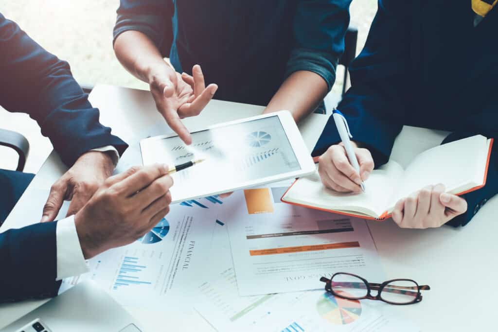 Business people in a meeting analysing data on a table device and taking notes in a notebook. Papers, glasses and a laptop is also on the desk.